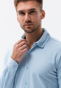 Polo de color azul claro, texturizado, con cuello abotonado. Botones en color a juego, mangas largas. El modelo toca el cuello con la mano.