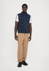 Navy padded vest with high collar, front zip, and two side pockets. Paired with beige cargo pants and black lace-up shoes.