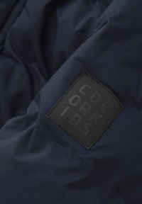 Marinblå vadderad jacka med en texturerad svart lapp som har den präglade texten "COLOR KIDS." Slät, syntetisk material; matt yta.