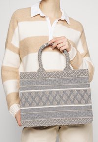 Szara, pleciona torba typu tote z misternymi, wzorzystymi akcentami. Posiada dwie solidne rączki oraz usztywniony, prostokątny kształt.