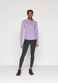 Ελαφρύ μοβ μακρυμάνικο τοπ με φερμουάρ έως το στήθος, συνδυασμένο με μαύρα κολάν, με εφαρμοστή σχεδίαση και επίπεδες ραφές για άνεση. Μαύρα αθλητικά παπούτσια ολοκληρώνουν το look.