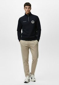 Navy sweatshirt with a high collar and zipper, featuring a circular patch. Beige tapered pants, gray and navy sneakers complete the look.