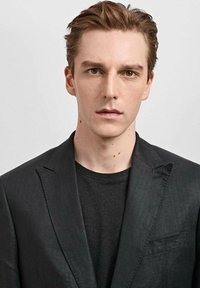 Schwarzer Blazer mit Reverskragen über einem schwarzen T-Shirt, aus glattem Stoff mit dezentem Muster. Der neutrale Hintergrund sorgt für Kontrast.
