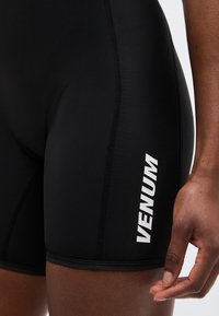 Κοντινό πλάνο του μηρού ενός ατόμου που φοράει στενά μαύρα αθλητικά σορτς με λευκή επιγραφή "VENUM" στο πλάι και ένα χαλαρό χέρι δίπλα του.