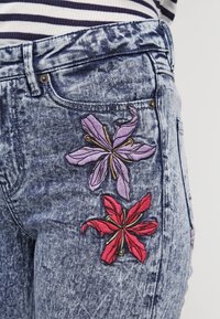 Dżinsy z widocznym niebieskim odcieniem, z różowym i fioletowym haftem kwiatowym na tylnej kieszeni, podkreślające skomplikowane detale wyszycia.