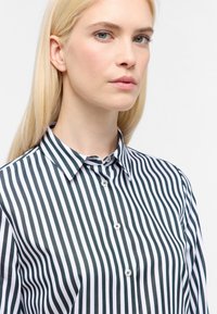 Camicia con bottoni a righe verticali bianche e nere, dotata di colletto a punta e chiusura con bottoni. Tessuto liscio con una leggera lucentezza.