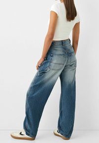 Vida jeans i denim med blekt blå tvätt och synliga sömmar, stora fickor och en lätt sliten textur, kombinerade med vit topp och sneakers.