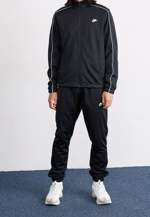 Persoon draagt een zwart Nike trainingspak met witte biezen langs mouwen en broek, gecombineerd met witte sportschoenen, staand op een blauw tapijt.