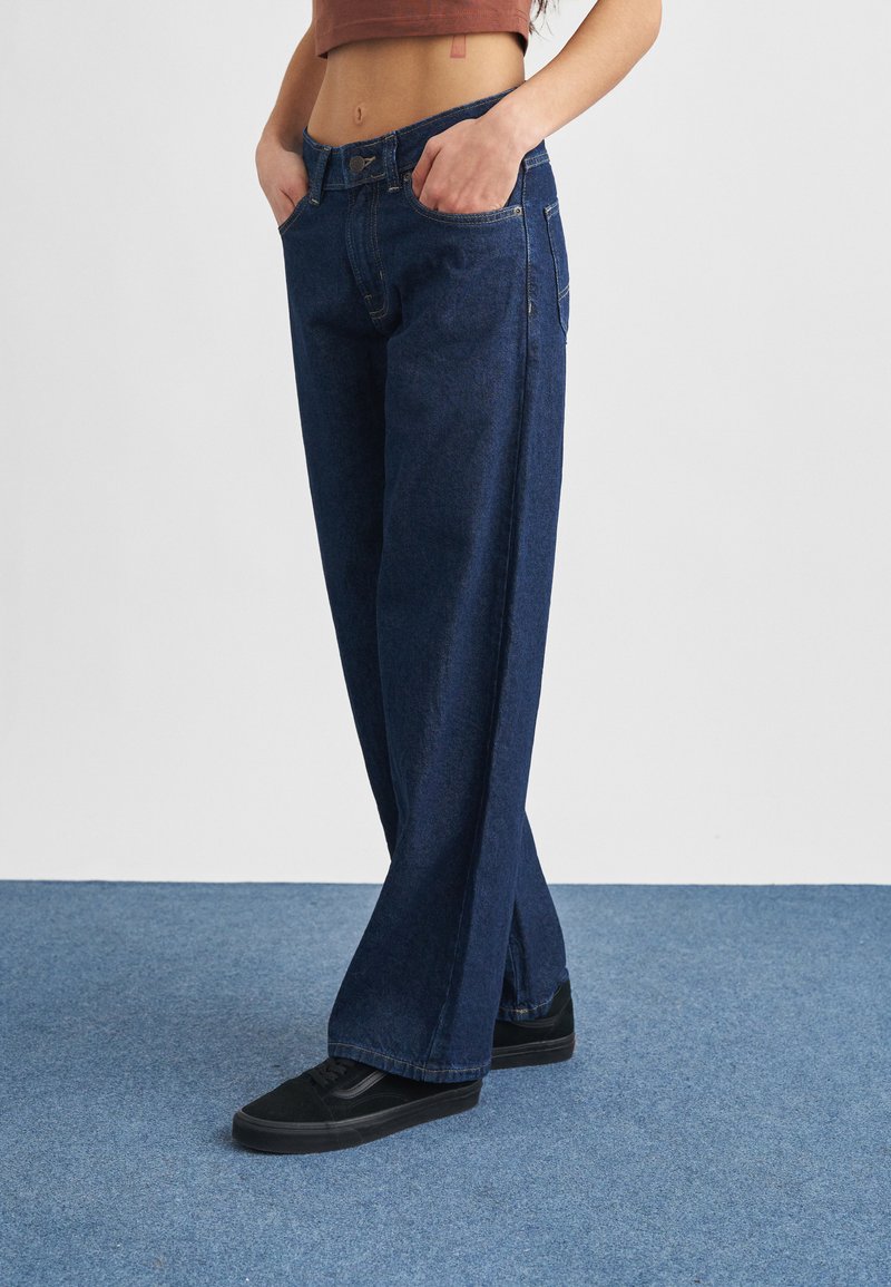 Personne portant un jean large bleu foncé et des baskets noires, debout les mains dans les poches sur un tapis bleu.