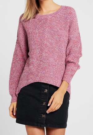 Femme portant un pull en tricot rose et une mini-jupe noire à boutons sur le devant, posant avec une main reposant près de sa taille.