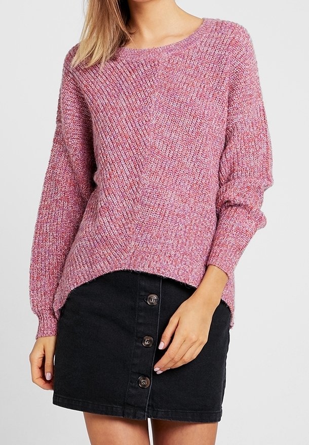 Femme portant un pull en tricot rose et une mini-jupe noire à boutons sur le devant, posant avec une main reposant près de sa taille.
