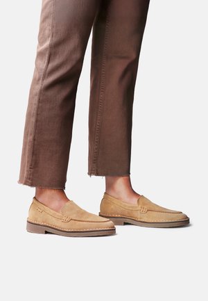Personne portant des mocassins en daim beige associés à un pantalon marron raccourci, debout sur un fond blanc.