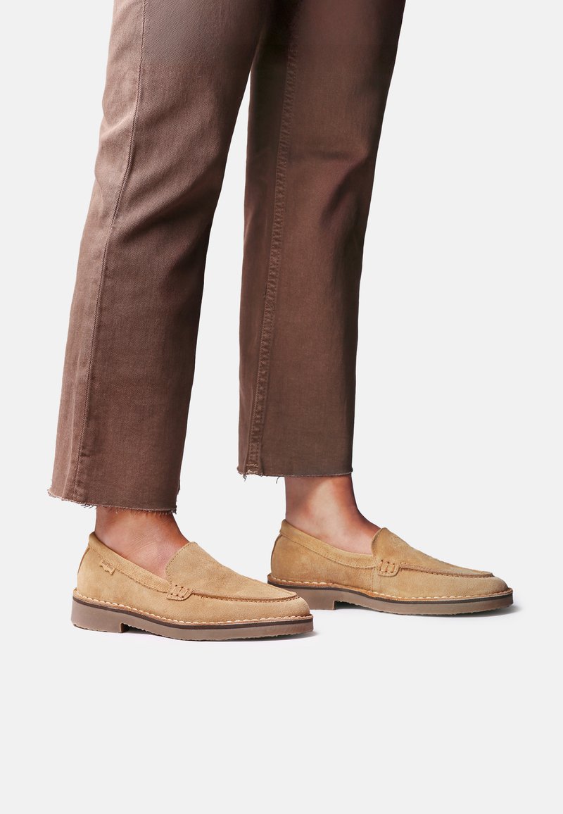 Personne portant des mocassins en daim beige associés à un pantalon marron raccourci, debout sur un fond blanc.