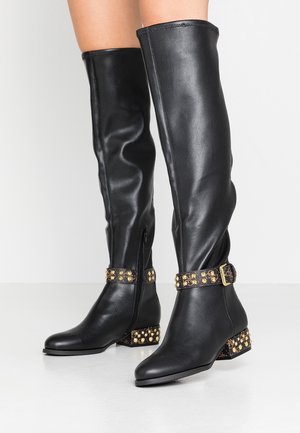 Zwarte kniehoge leren laarzen met goudkleurige gespen en hakken, gedragen op blote benen tegen een witte achtergrond.