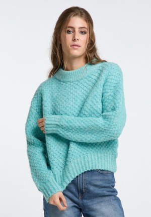 Pulover texturat de culoare albastru deschis, cu un model gros de tricot, guler rotund și margine ribbată. Asortat cu blugi din denim.
