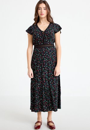 Jeune femme portant une robe midi noire à motifs floraux avec des manches courtes et des chaussures plates rouges, se tenant devant un fond blanc uni.