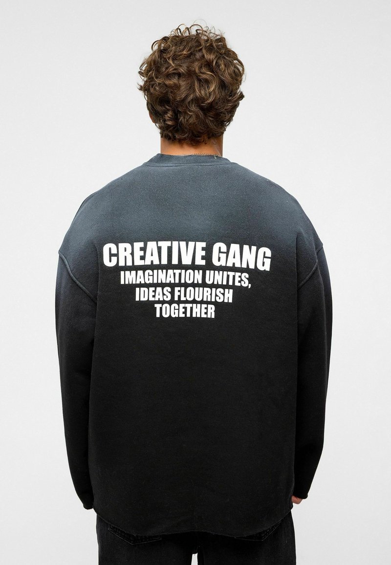 Zwarte sweatshirt met een ronde hals en een drop-schouder ontwerp. De achterkant heeft gedurfde witte tekst: "CREATIEVE BAND IMAGINATIE VERBINDT, IDEEËN GEVEN SAMEN GROEI."