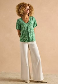 Zaļš blūze ar rakstu, īsām piedurknēm, V-veida kakla izgriezumu un saburzītu detaļu priekšā, pārī ar platām baltām biksēm un bēšām kurpēm.