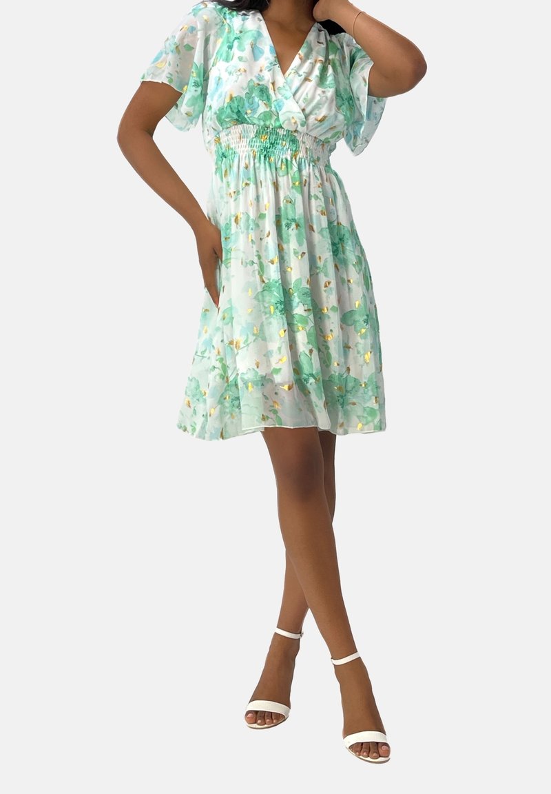 Femme portant une robe blanche mi-longue à imprimé floral vert et des accents dorés, associée à des sandales blanches à bride à la cheville, posant avec une main sur la hanche.