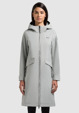 Nő világosszürke, térdig érő, kapucnis kabátban, elöl cipzárral és fedett zsebekkel, sima háttér előtt áll.