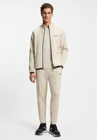 Beige athletic tracksuit featuring a zip-up jacket with black trim and logo, paired with tapered pants that have zippered ankle details.