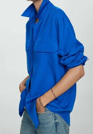 Personne portant une chemise ample bleu vif à boutons avec manches retroussées et un jean bleu clair, les mains dans les poches avant.