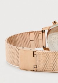 Rose gold mesh watch band with a smooth texture, featuring a secure clasp and a detailed patterned weave. Back shows the watch's inner mechanics.