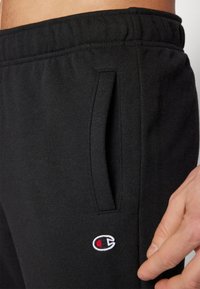 Svarta sweatpants gjorda av mjukt material med elastiskt midjeband, sidofickor och en liten broderad logotyp i rött och vitt.