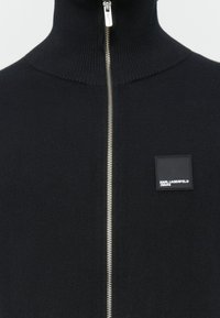 Melnā augsta kakla zip-up džemperis ar sudraba rāvējslēdzēju un nelielu kvadrātu "Karl Lagerfeld Jeans" logo marķējumu uz krūtīm.