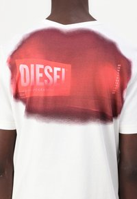 Camiseta de algodón blanca con un gráfico de pintura en spray roja que presenta el logotipo "DIESEL" y texto en la parte posterior. Textura suave y corte casual.