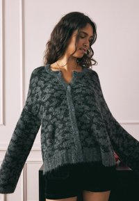 Sivi oversized kardigan u pahuljastom, teksturiranom pletenju s uzorkom leoparda i suptilnim sjajem. Ima okrugli izrez i kopčanje na gumbe.