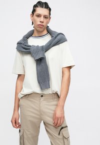 Γκρι πλεκτό πουλόβερ δεμένο γύρω από τον λαιμό, φορεμένο πάνω από λευκό μπλουζάκι. Μπεζ παντελόνι cargo με πλαϊνές τσέπες και υφή.