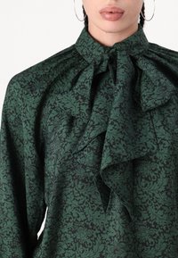 Blusa floreale verde con colletto alto e grande fiocco. Il tessuto presenta un motivo testurizzato con foglie e fiori verde scuro su uno sfondo nero.