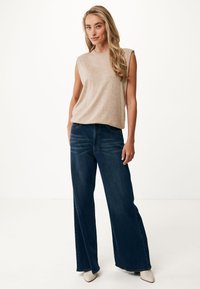 Top beige senza maniche con scollo rotondo, abbinato a jeans a gamba larga blu scuro. L'outfit è completato da scarpe bianche a punta.