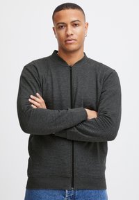 Chaqueta bomber de cremallera en gris oscuro con textura acanalada, cuello alto y mangas largas. Presenta una cremallera frontal y una cintura ajustada.