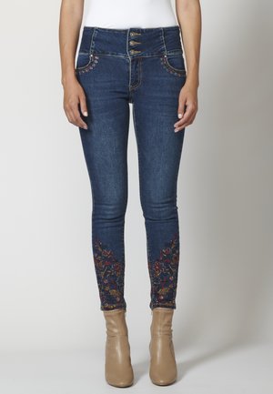 Dunkelblaue Slim-Fit-Jeans mit hohem Bund und dreifachem Knopfverschluss, florale Stickerei an den unteren Hosenbeinen, kombiniert mit beigen Stiefeletten und weißem Top.