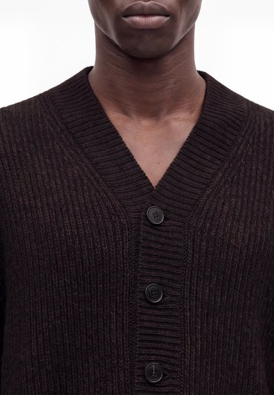 Gros plan d'une personne portant un cardigan en maille côtelée marron foncé avec de gros boutons noirs, montrant le cou et le haut de la poitrine.