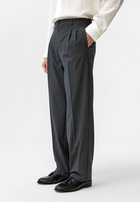 Grijze kostuumbroek met fijne strepen, wijd uitlopend, met plooien aan de voorkant en zijzakken. Gepaard met zwarte leren schoenen en een witte blouse met lange mouwen.