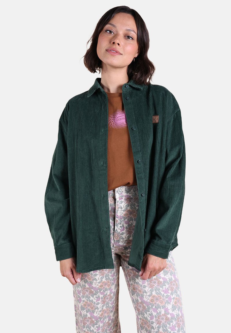 Chemise en velours côtelé vert avec fermeture à boutons, tissu texturé, modèle portant un t-shirt graphique marron et un pantalon à motif floral.