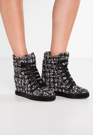 Ben iført høje kile-sneakers med sort og hvid tweedstof og brede sorte båndsnører på en hvid gulv.