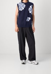 Marineblauer ärmelloser Pullover mit weißem Blumenmuster, kombiniert mit schwarzen gestreiften Hosen. Das Outfit wird mit silbernen Sportschuhen abgerundet.