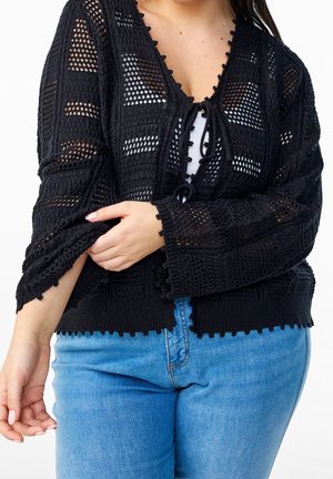 Femme portant un cardigan noir à mailles ajourées noué à l'avant sur un haut blanc et un jean bleu, ajustant la manche du cardigan sur son bras.