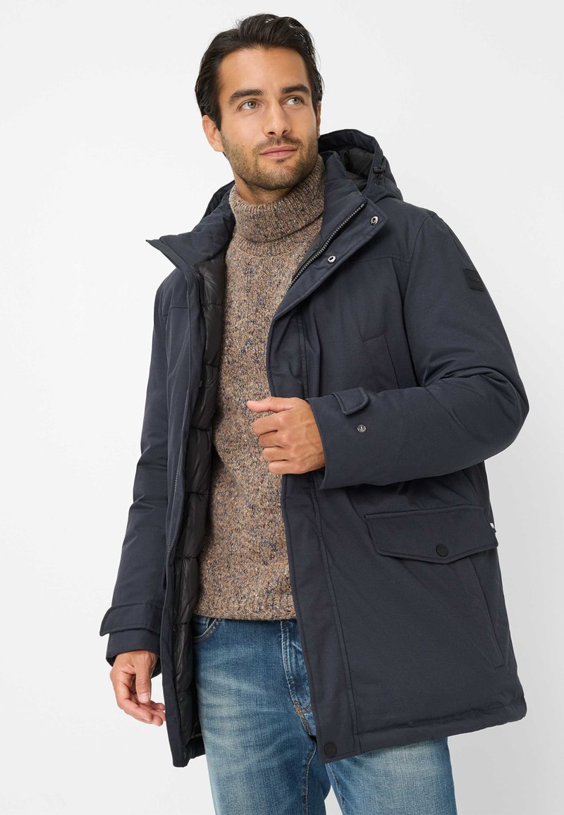 Man in een marineblauwe winterjas over een bruine coltrui en blauwe jeans, staand tegen een effen witte achtergrond.