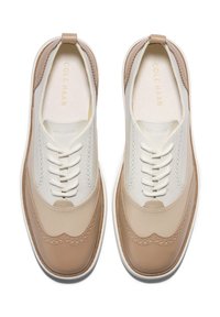 Pár béžových a bílých kožených wingtip tenisek s bílými tkaničkami, bílými mezipodešvemi a značkováním "Cole Haan" na vnitřních podešvích.