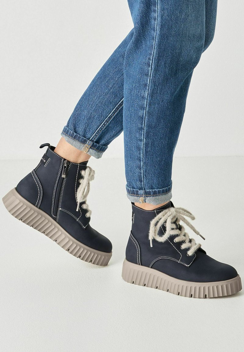 Personne portant un jean bleu retroussé et des bottes montantes bleu marine avec des semelles épaisses beige et des lacets blancs duveteux.