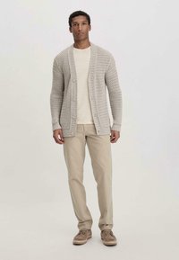 Hellgrauer gestrickter Cardigan mit einem strukturierten Muster, ausgestattet mit Knöpfen und einem lässigen Schnitt, kombiniert mit einem hellen Hemd und beigen Hosen.