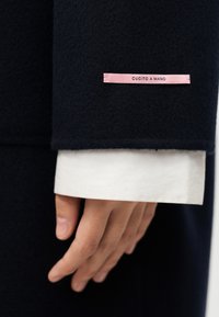 Μανίκι παλτού από ναυτικό μαλλί με ροζ σατέν ετικέτα που γράφει «CUCITO A MANO». Λευκό μανίκι διακρίνεται από κάτω, προσθέτοντας αντίθεση.