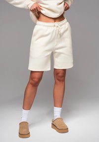 Shorts de survêtement couleur crème en tissu doux, avec une taille élastique et un cordon de serrage, et un petit logo néon en accent.