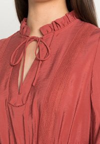 Blusa color ruggine con colletto arricciato, scollatura a V e dettaglio con fiocco. Presenta un tessuto testurizzato e accenti di pizzo verticale sui lati.