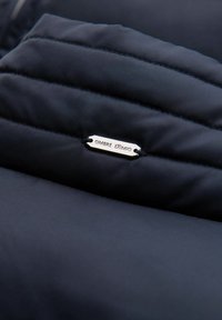 Marineblå pufferjakke-sektion med quiltet tekstur, der har et metallisk label med teksten "OMBRE EXTIMO" og forstærkede sømdetaljer.